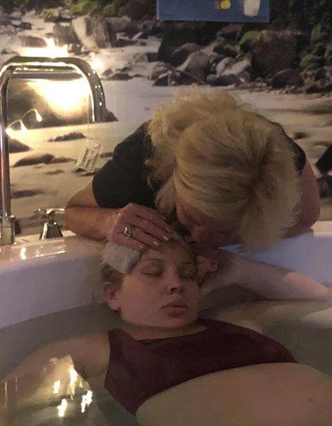 Pregnant mum lying in birth pool with cloth on her forehead by birth support