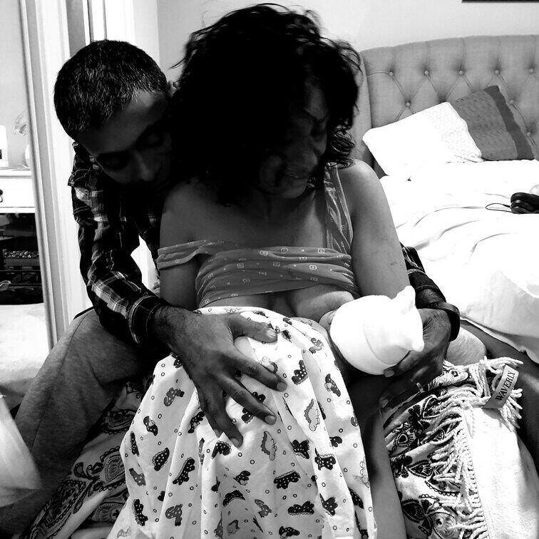 Mum sitting on bed holding newborn baby with partner by her side