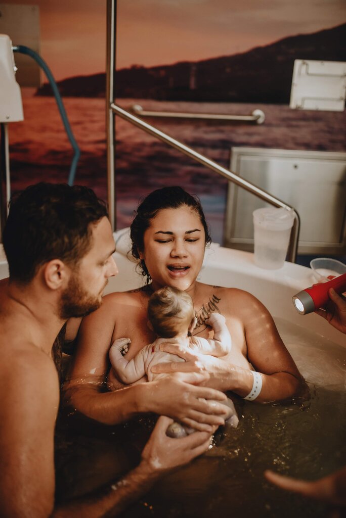 Mum holding her newborn baby on her chest in birth pool with partner by her side