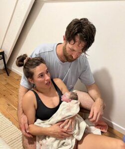 Mum sitting on floor at home with newborn baby in her arms and partner by her side
